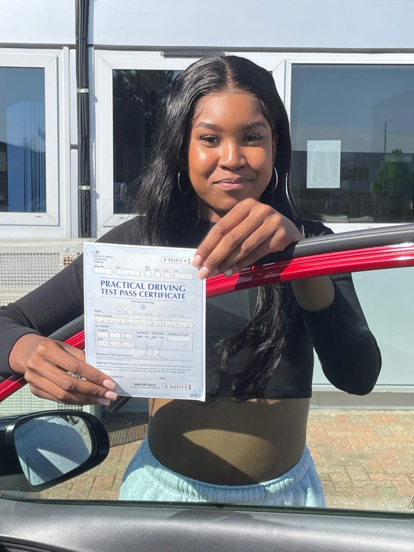 Learner holding her practical driving test pass certificate beside the red Hallwood Training Services car