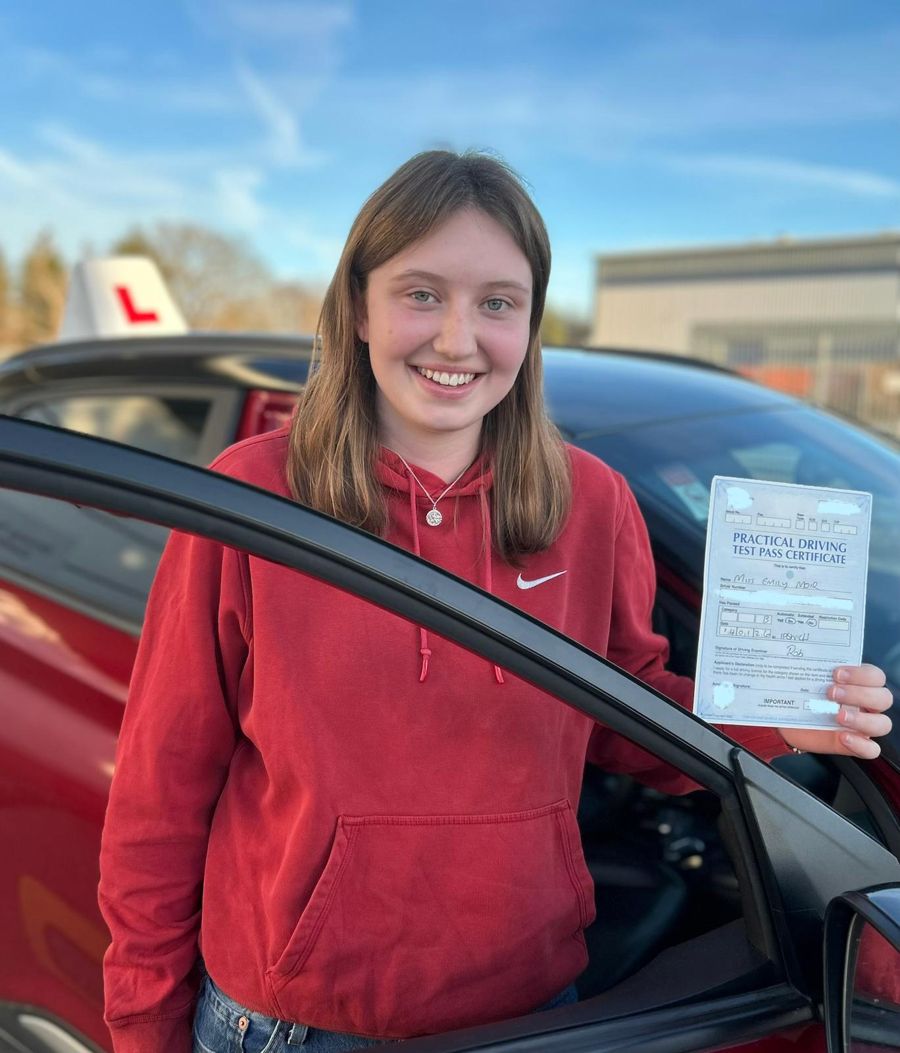 Smiling learner in a red hoodie holding her pass certificate at the open door of the Hallwood Training Services car
