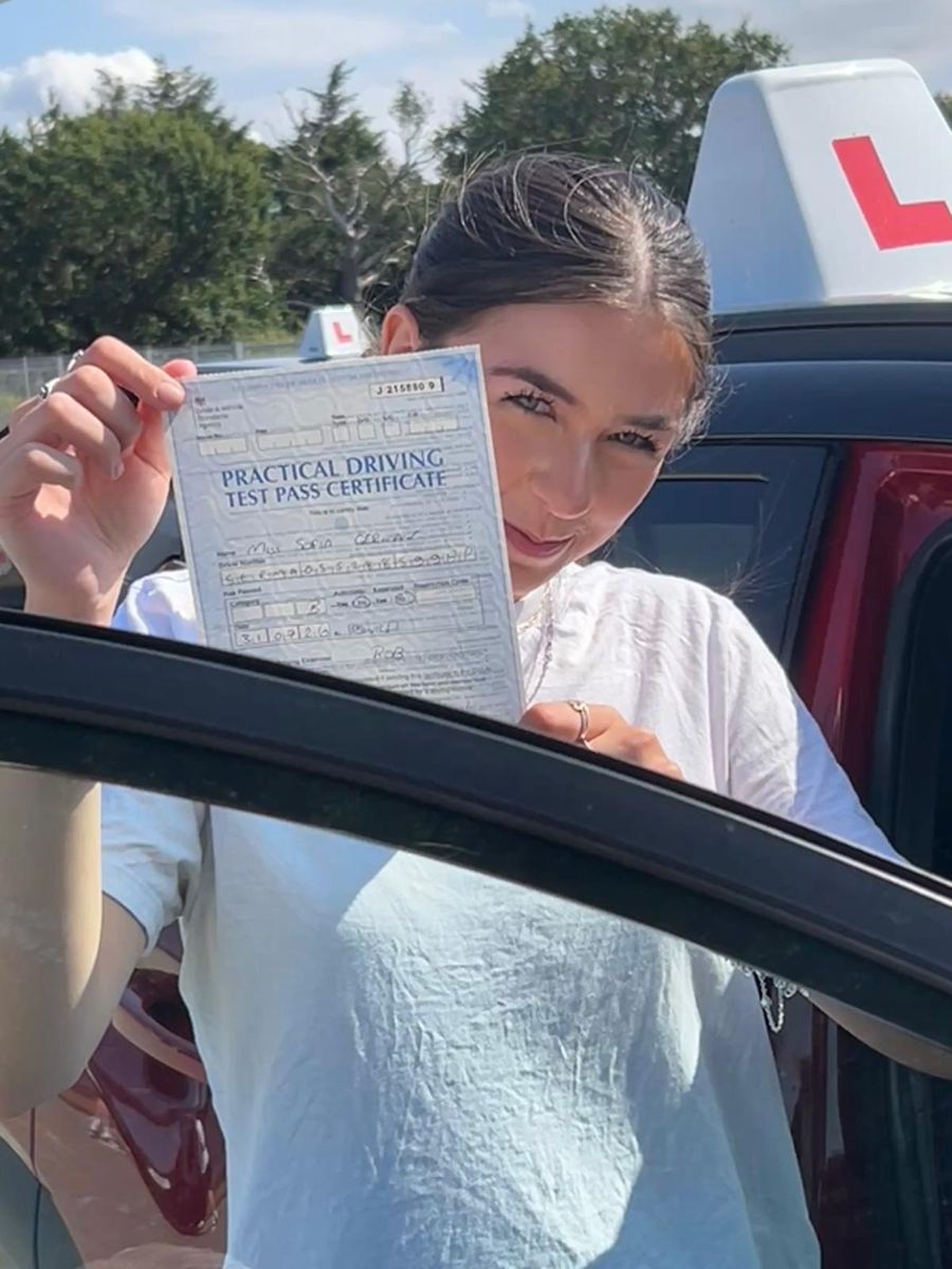 New driver holding her practical driving test pass certificate up to the camera beside the Hallwood Training Services car