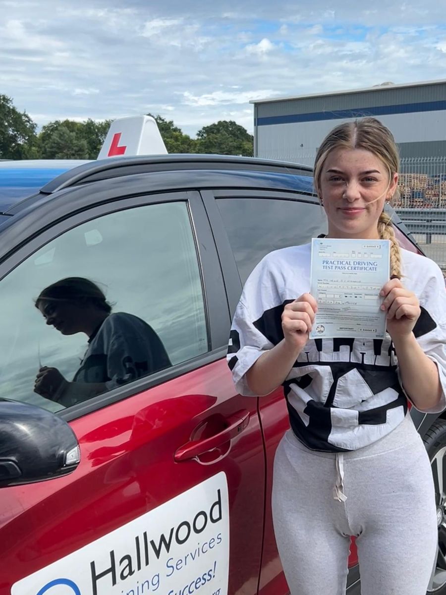 Pupil holding her pass certificate beside the red Hallwood Training Services car with its branded door sign