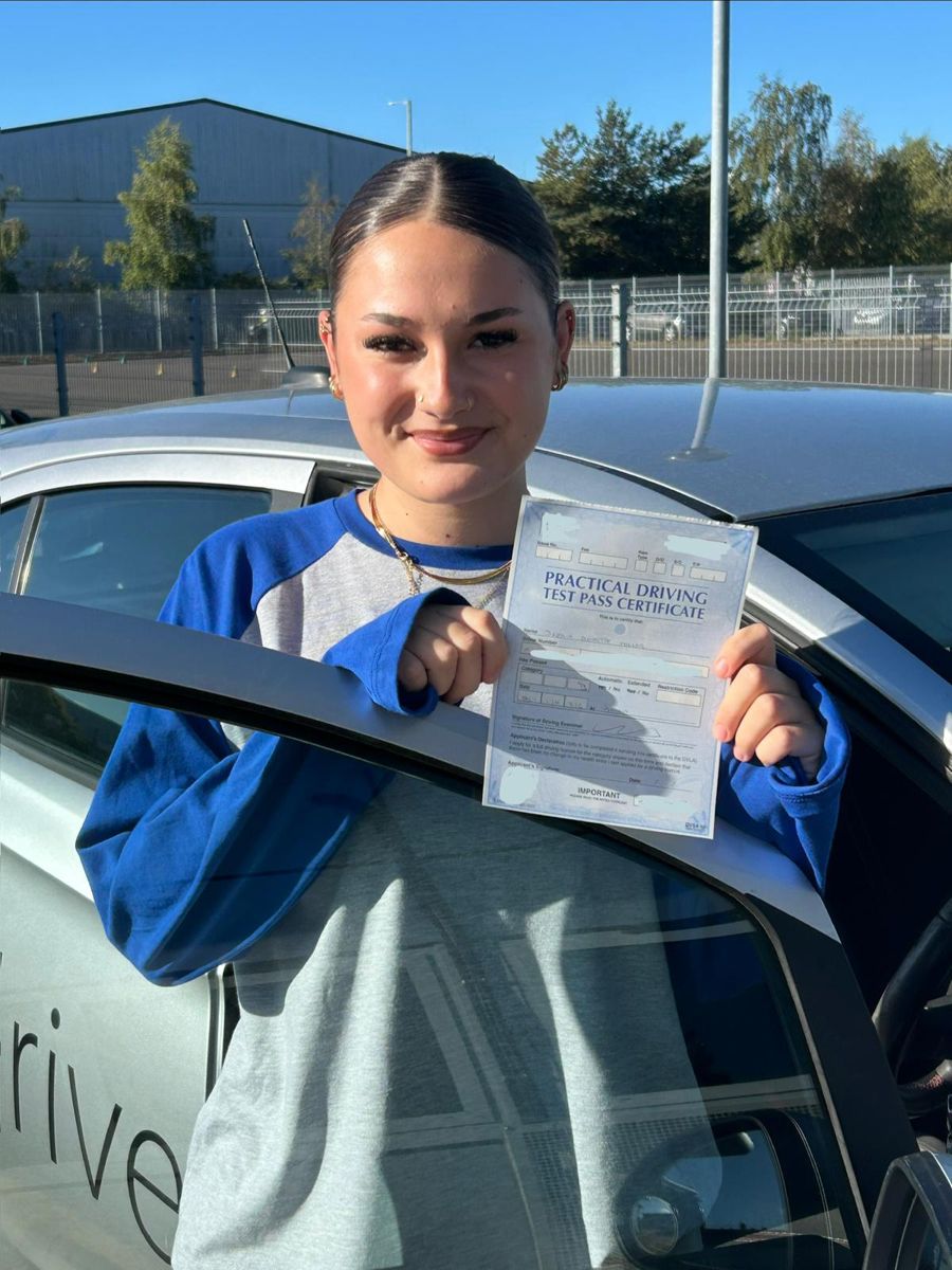 Young driver in a blue top holding up her driving test pass certificate by the car door